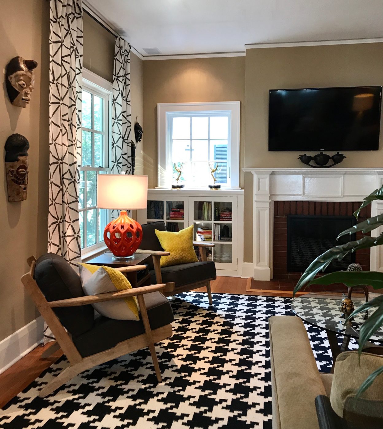 Mid-century modern lounge chairs sit atop a black and white hounds-tooth rug. Canary yellow pillows adorn chairs. Nestled in between chairs is a side table with a orange mid-century modern lamp. Displayed on the wall are masks from the homeowners collectibles.